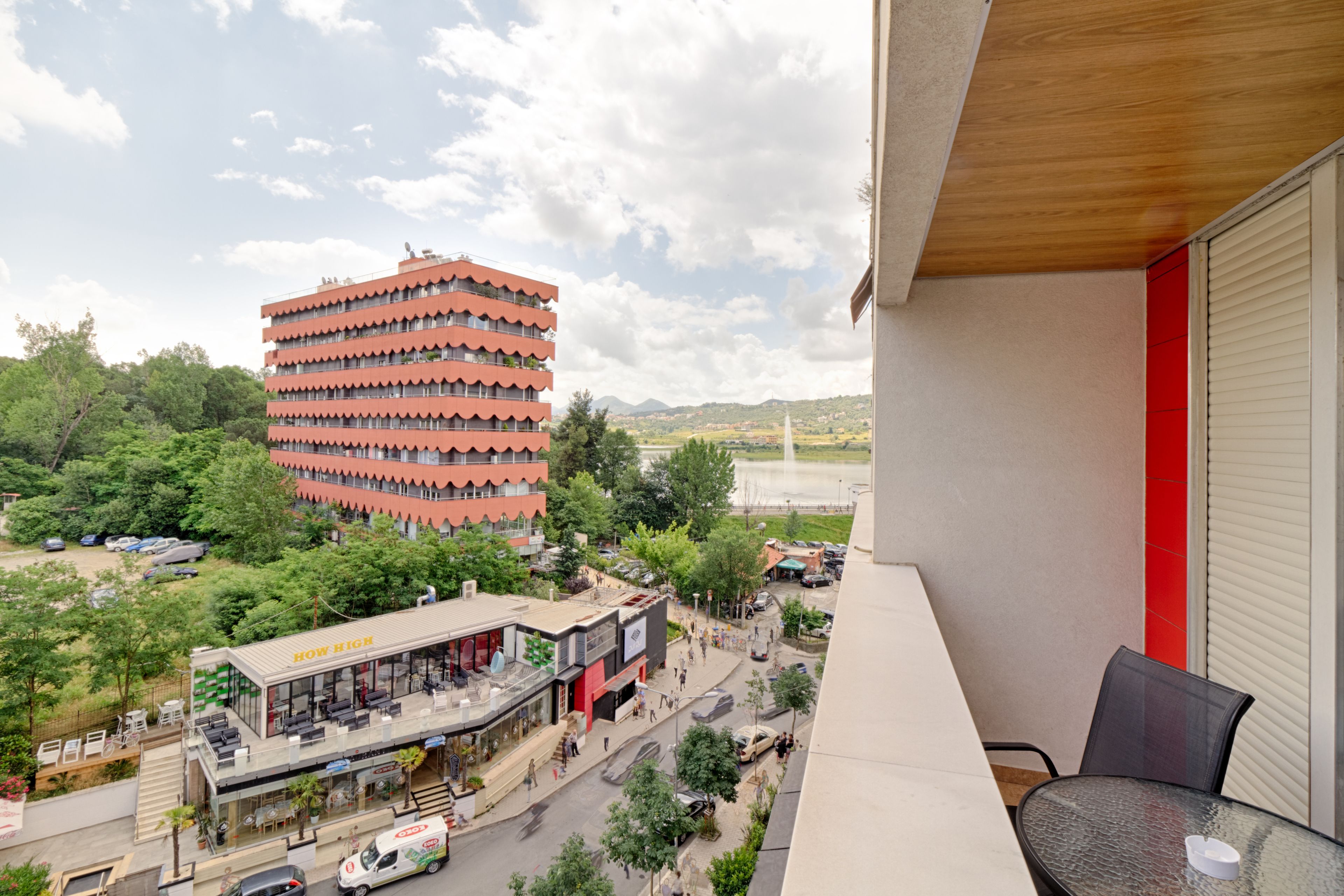 Un balcón con vista a un edificio de varios pisos, un restaurante y una calle con vehículos.