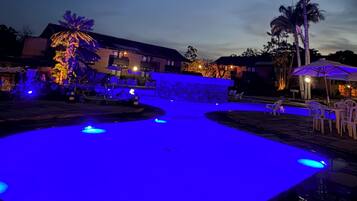 Zona de piscina con iluminación azul, palmeras y mobiliario exterior.
