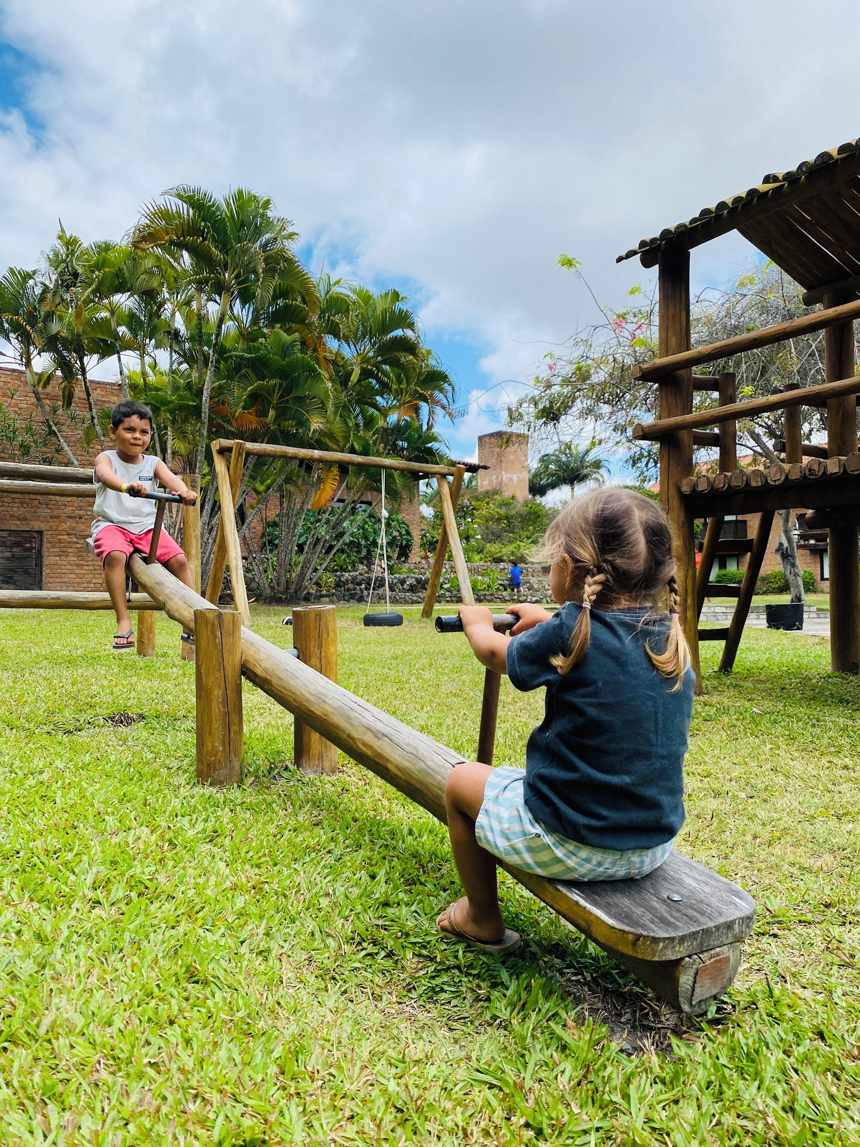 Dos niños jugando en un sube y baja en un espacio exterior con césped, estructuras de madera y palmeras al fondo.