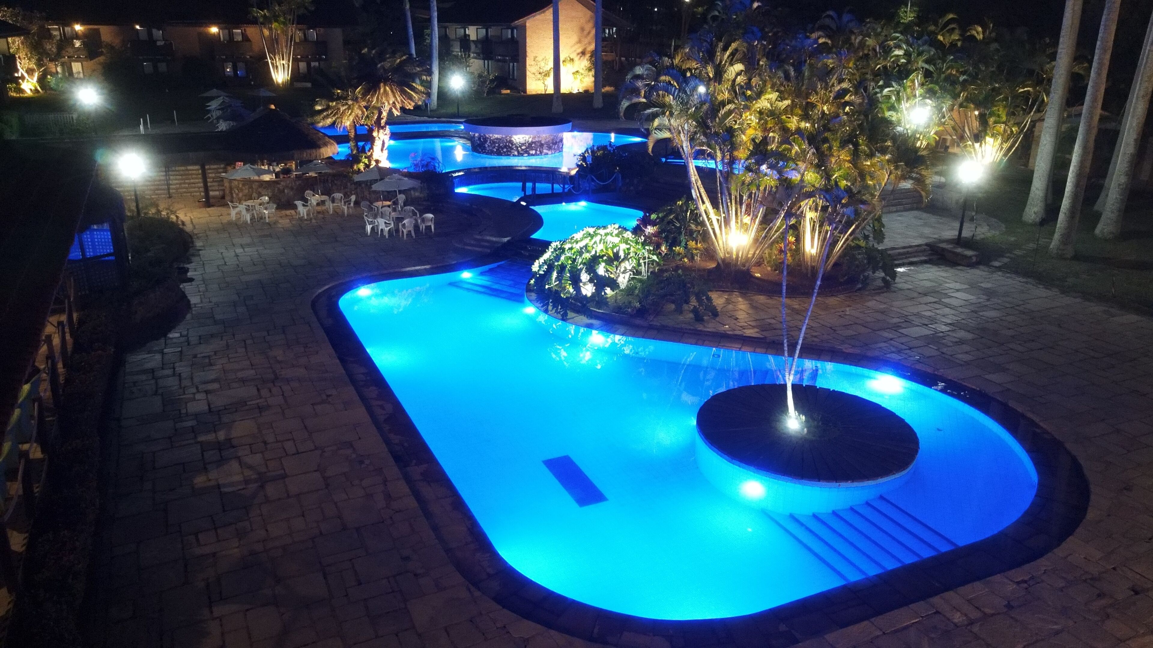 Une piscine nocturne dont le bord est illuminé de lumière bleue, entourée de palmiers et bordée d’un chemin en briques.