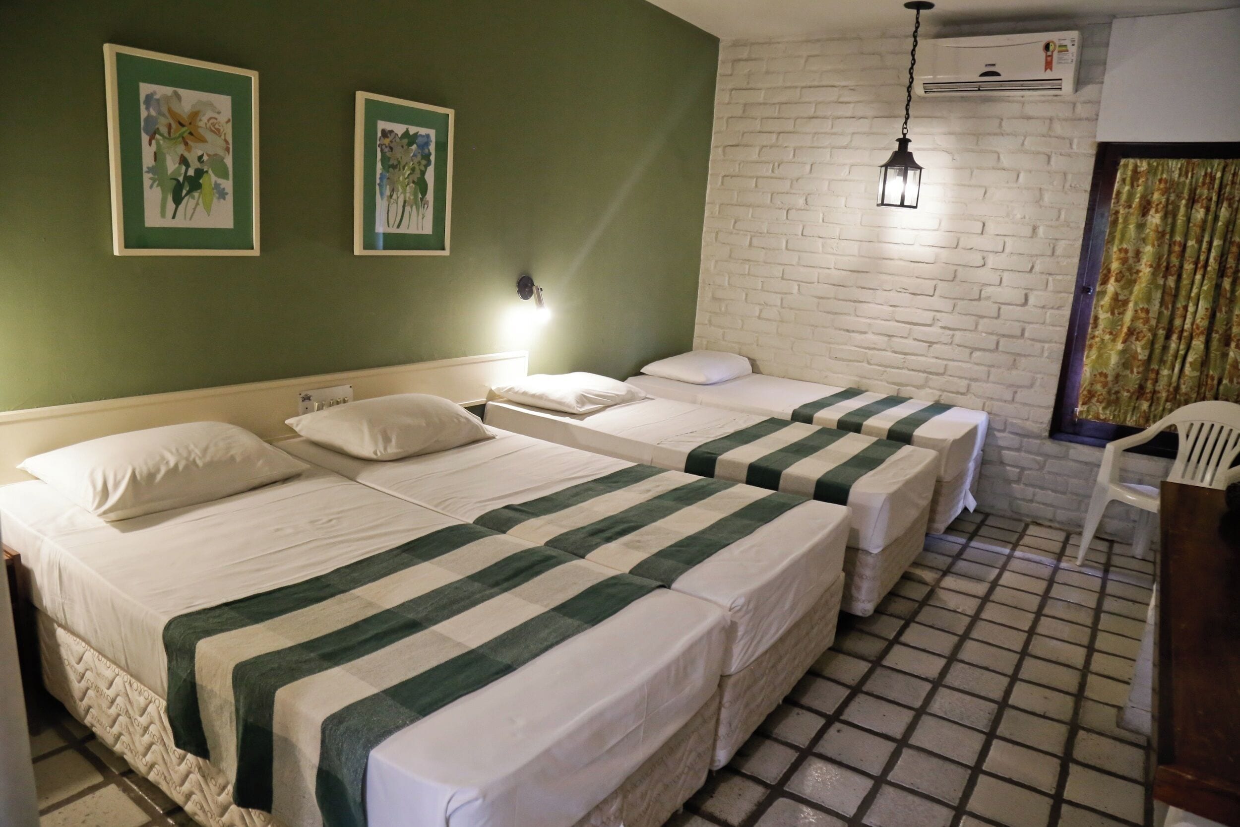 Ein Zimmer mit zwei Betten, jeweils mit einer grün gestreiften Bettdecke, weißen Kissen und einem weißen Kopfteil. Eine weiße Backsteinwand, eine Pendelleuchte und ein weißer Stuhl sind ebenfalls zu sehen.