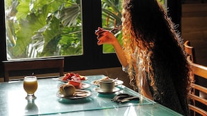 Una persona sentada a una mesa con un vaso de jugo, platos de comida y una taza. La mesa está cerca de una ventana con vistas a la vegetación.