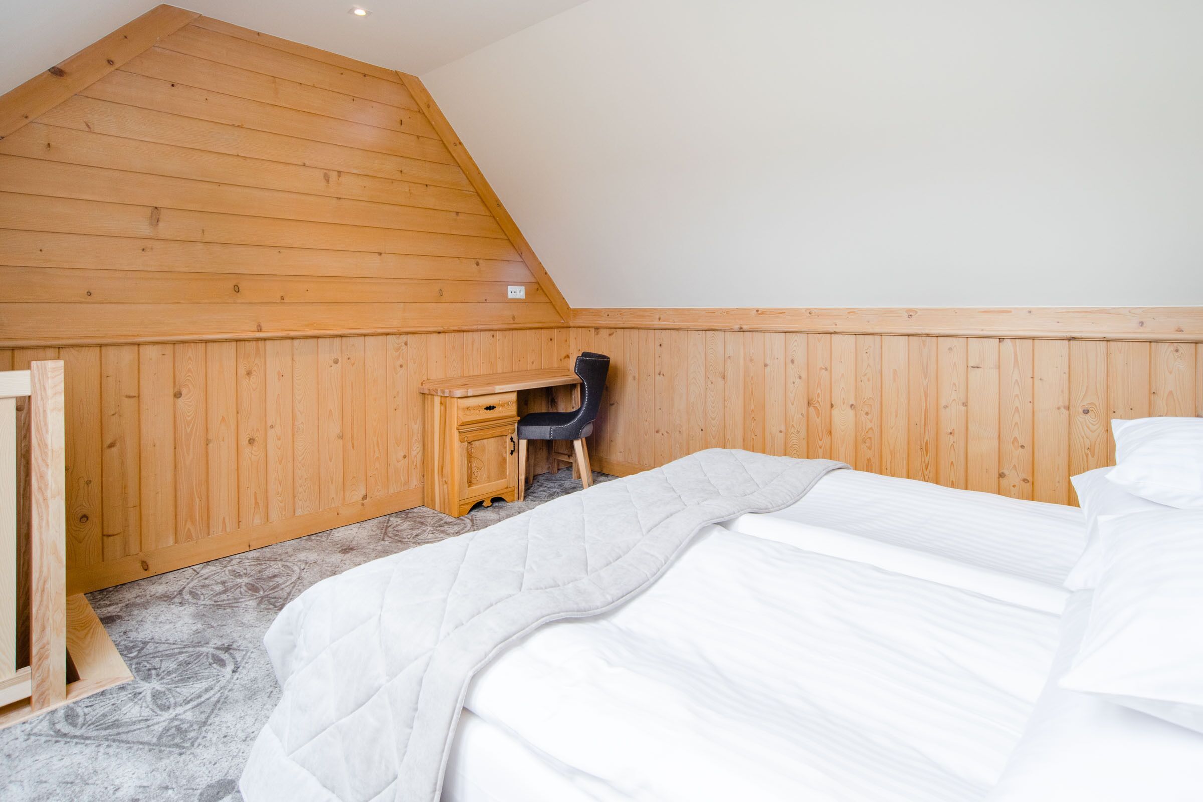 Una camera da letto con soffitto spiovente, pareti in legno, un letto con lenzuola bianche, una scrivania con una sedia e una piccola finestra.