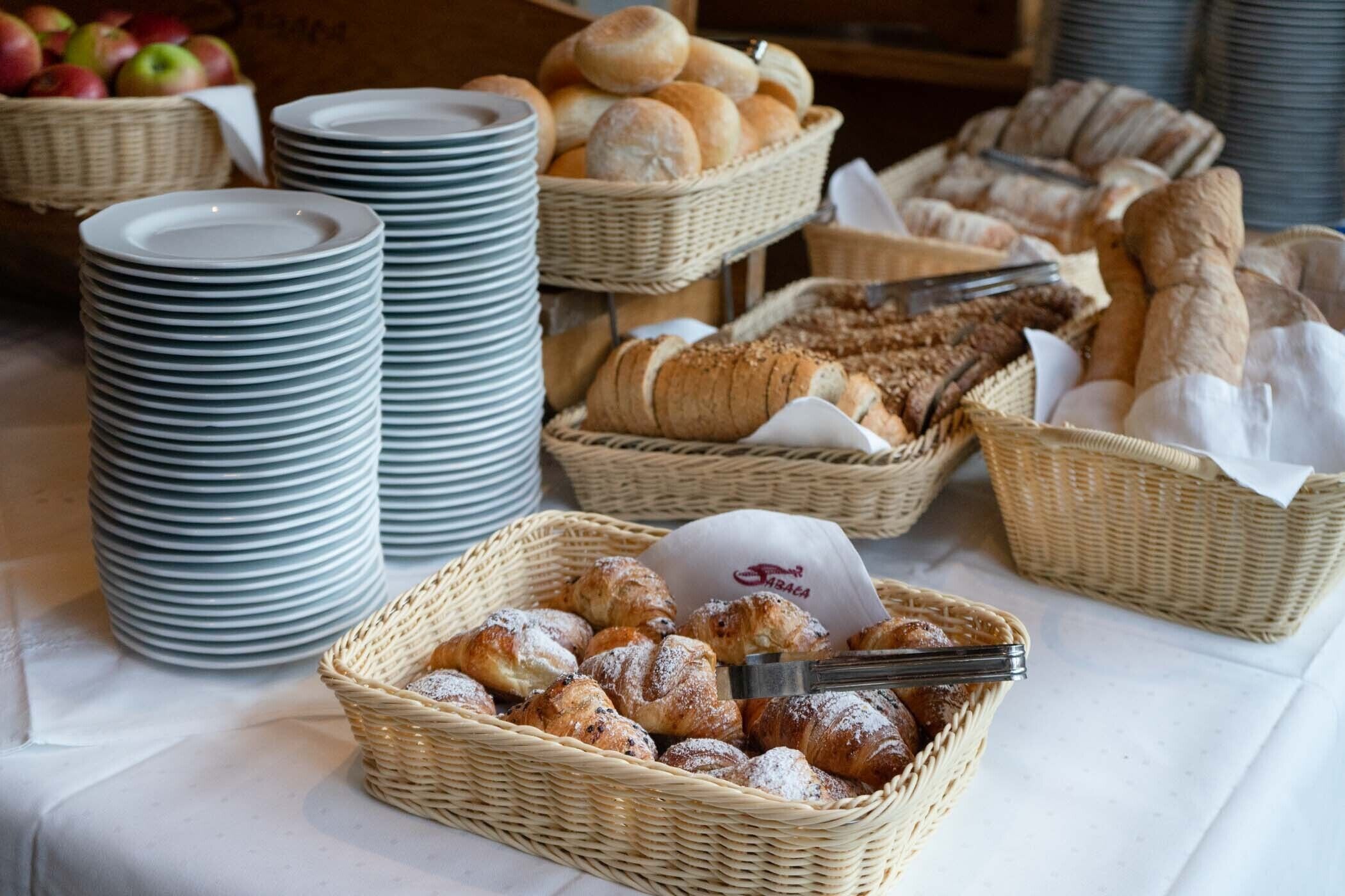 A buffet table with stacks of white plates, baskets of bread, and baskets of pastries.