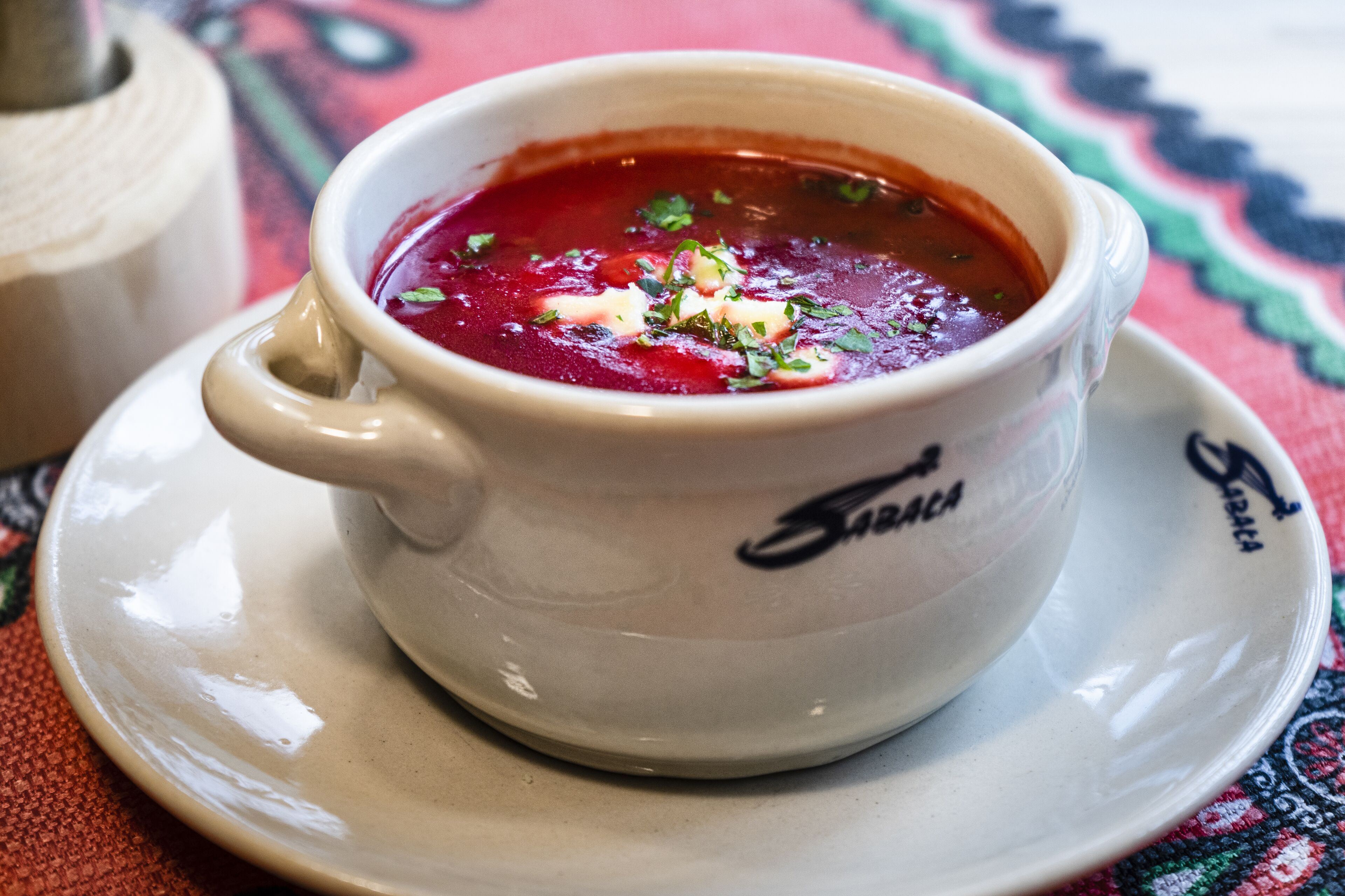 Una taza de cerámica blanca con asa, llena de sopa roja, sobre un plato hondo blanco.