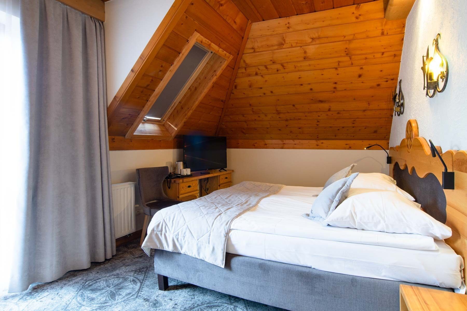 Un dormitorio con techo inclinado, cama con ropa de cama blanca, una silla, escritorio con televisor y ventana con cortinas.