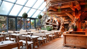 A restaurant with wooden tables and benches, a stone wall with a wooden wheel, and hanging greenery. - Hotel Sabala (Zakopane)