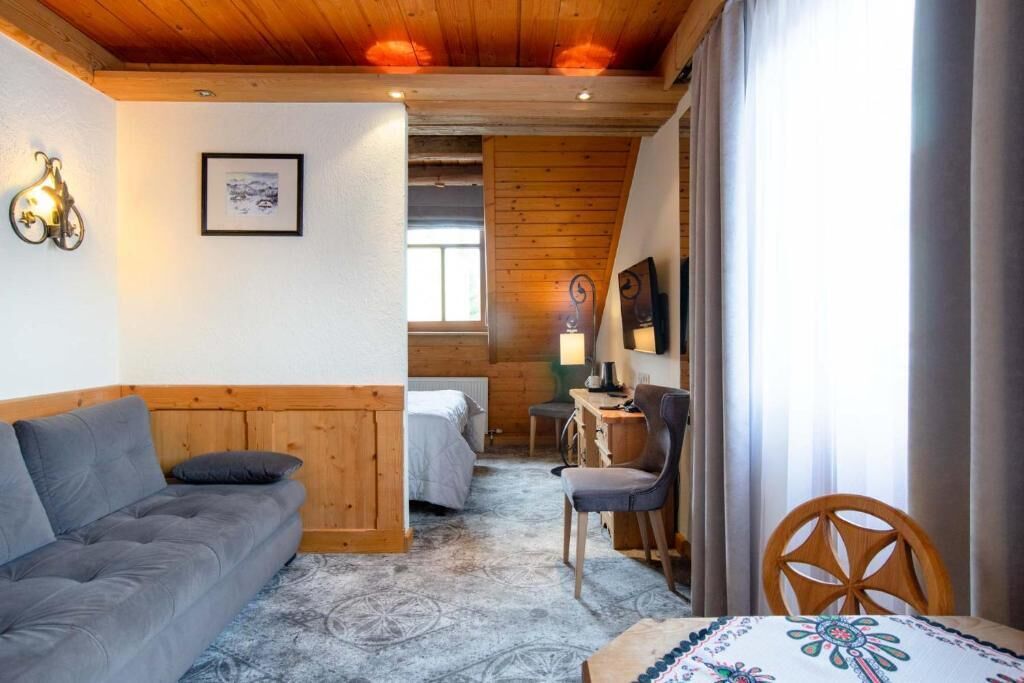 A cozy room with a wooden ceiling, a grey sofa, a bed, a desk with a chair, and a table with a patterned tablecloth.