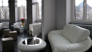 A modern living room with a white sofa, a round coffee table, and a glass-top side table. There are two white armchairs and a small glass table with a vase of flowers. The room has large windows with a view of trees and buildings outside. - Hôtel Alerion (Metz)