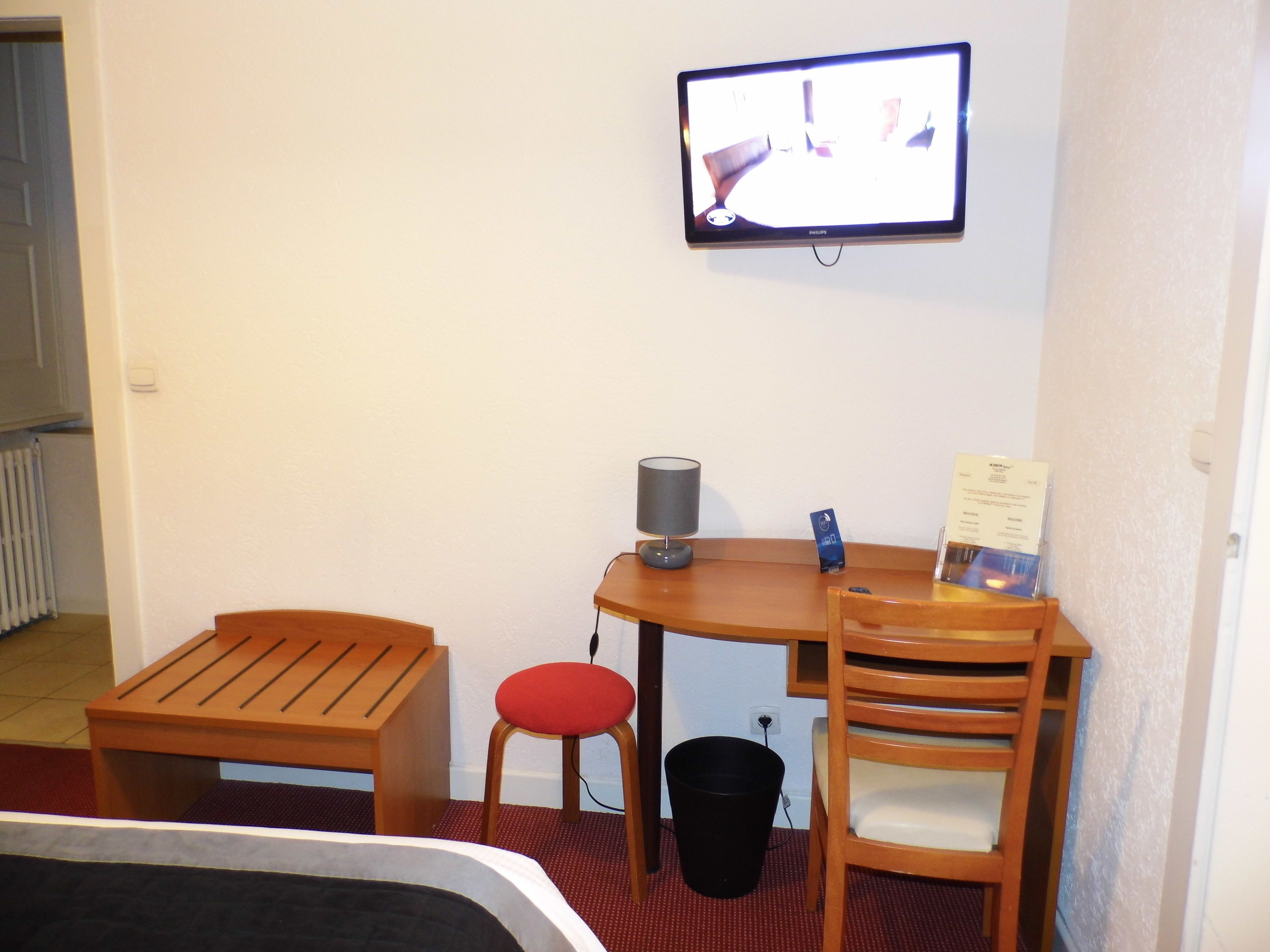Een kleine hotelkamer met een bed, een houten bureau, een rode stoel, een zwarte prullenbak en een aan de muur bevestigde televisie.