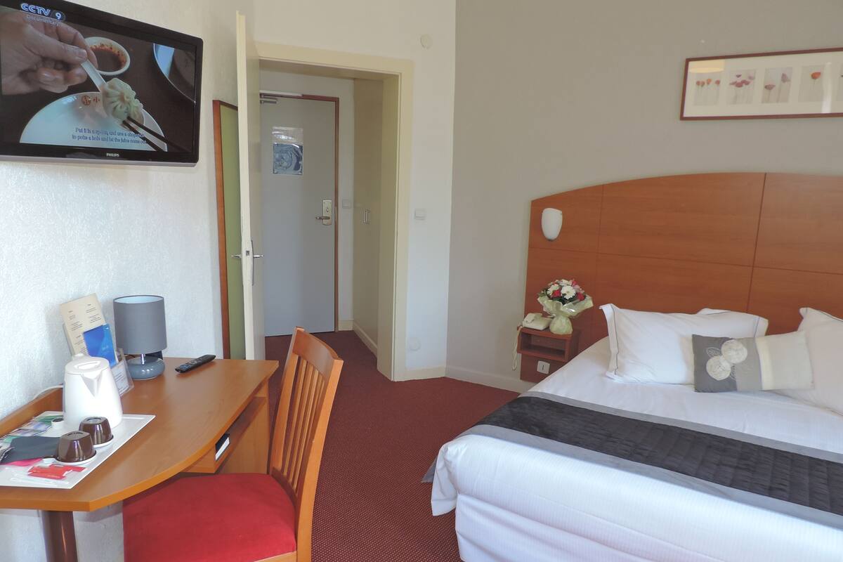 Ein Hotelzimmer mit Bett, Schreibtisch mit Stuhl, Fernseher, Nachttisch mit Telefon und Blumen sowie einem gerahmten Bild an der Wand.