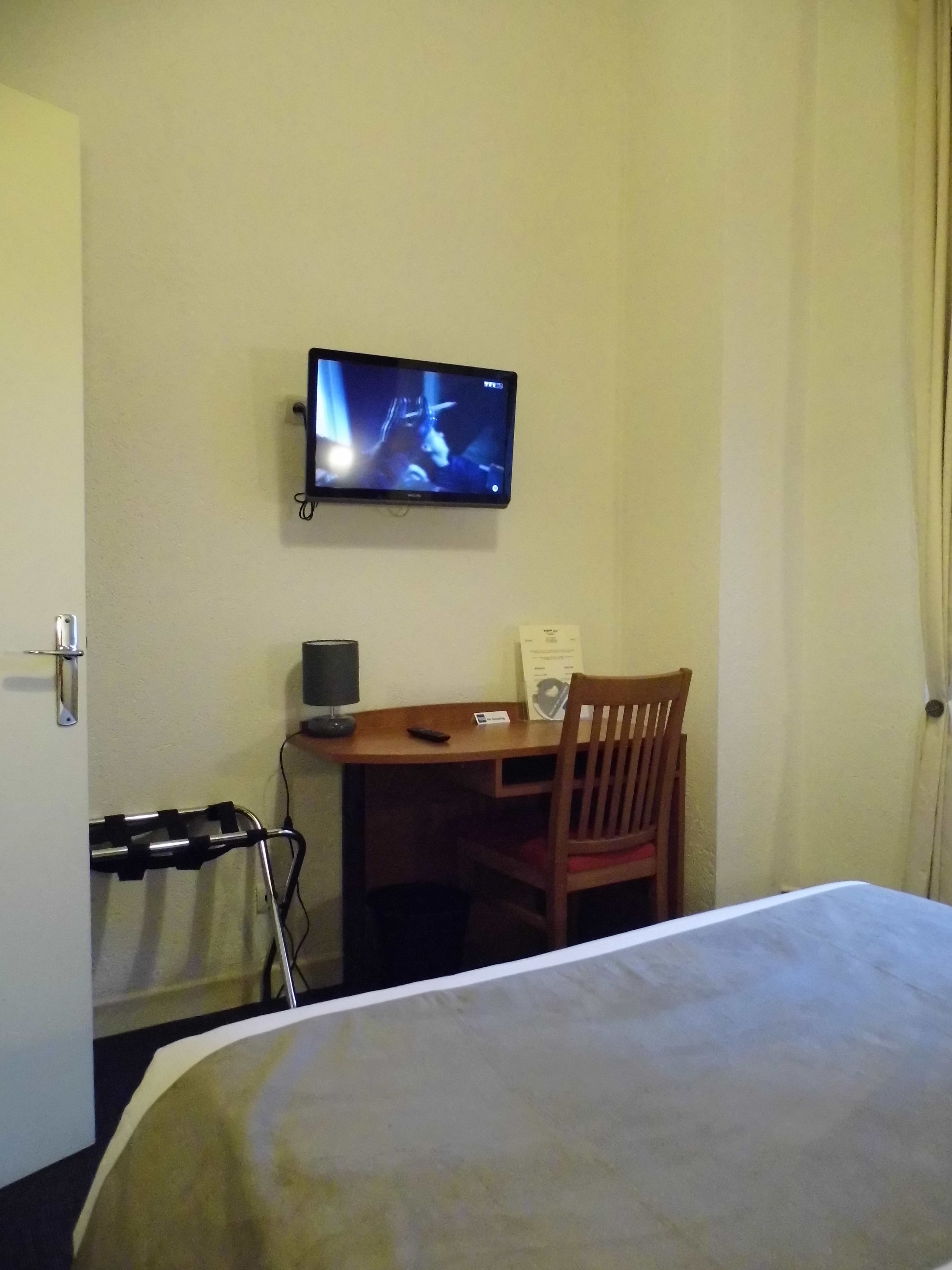 A hotel room with a bed, a desk with a chair, a television mounted on the wall, and a wheelchair.