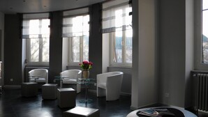 A modern living room with a curved layout, featuring a white sofa, a glass coffee table, and a white armchair. A vase with flowers is placed on the coffee table. - Hôtel Alerion (Metz)