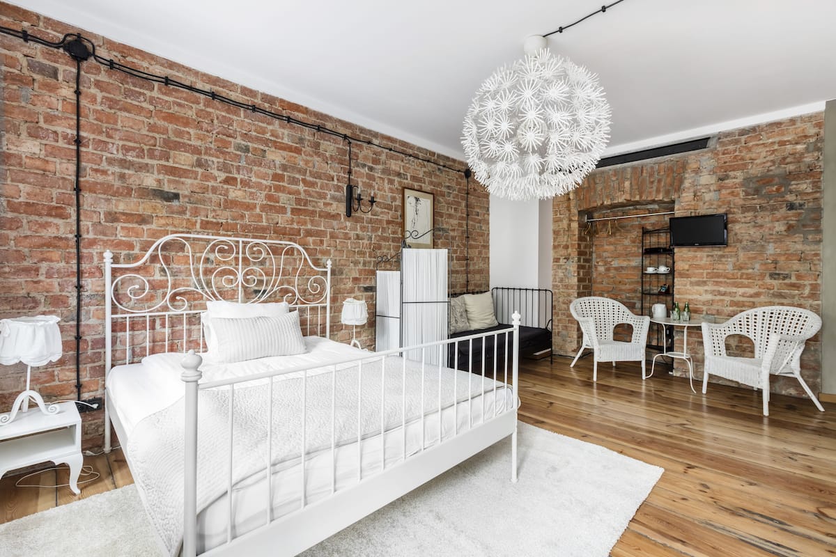 A bedroom with a white metal bed, white wicker chairs, a white chandelier, and a brick wall.