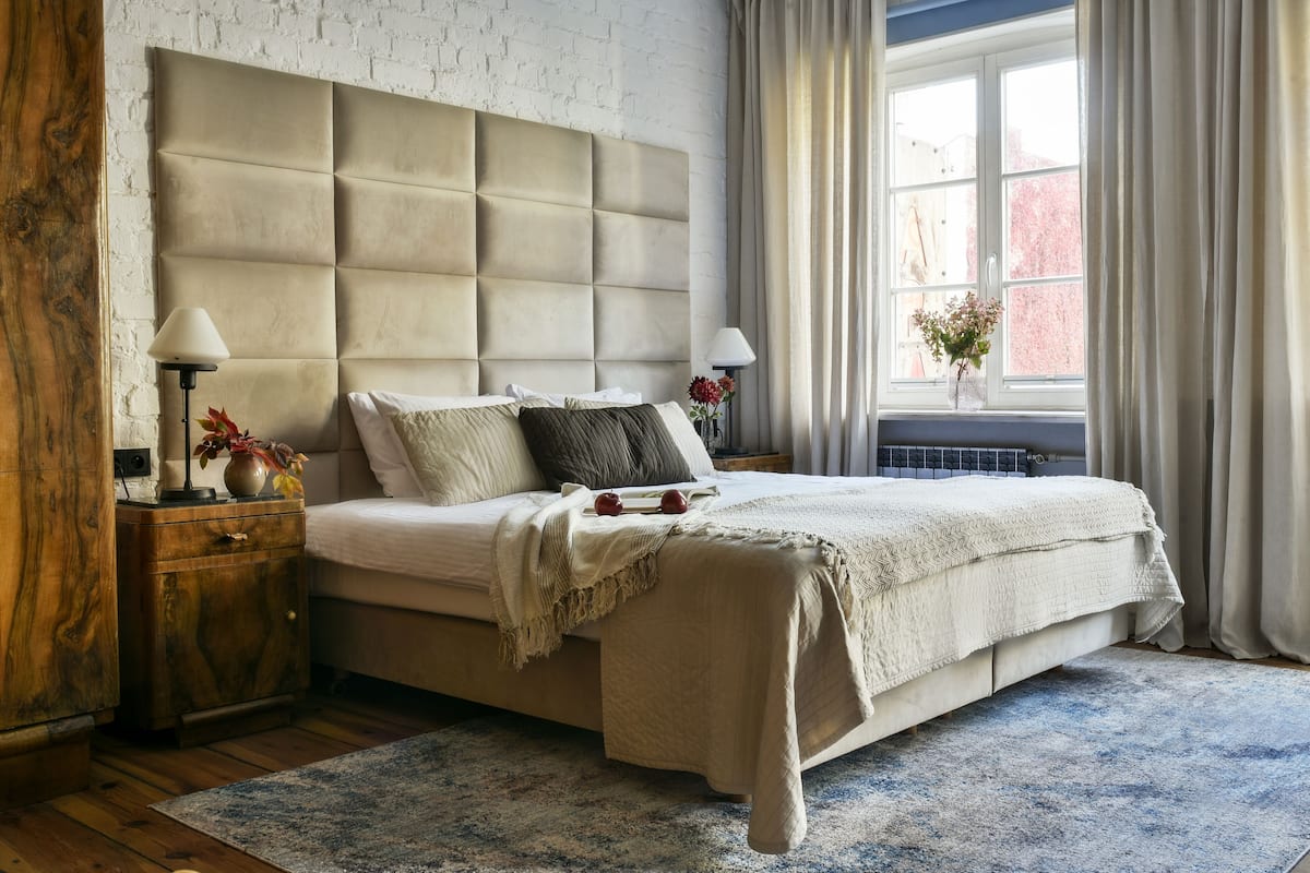 A bedroom with a tufted headboard, a large window with curtains, a bedside table with a lamp, and a decorative vase.