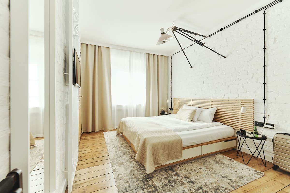 A modern bedroom with a wooden floor, a large bed with a wooden headboard, a bedside table with a lamp, and a suitcase on the floor.