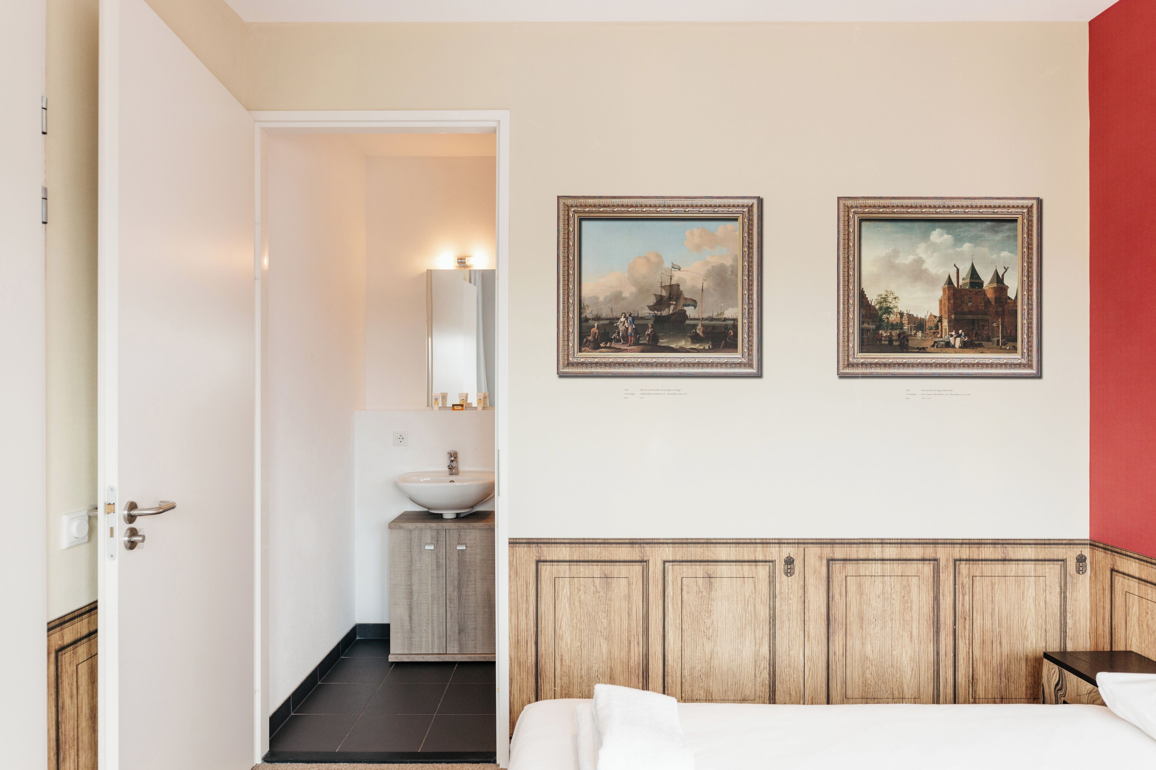 Une chambre d’hôtel avec une tête de lit en bois, un lit, une table de chevet, une commode, un miroir fixé au mur, un lavabo et deux tableaux encadrés accrochés au mur.