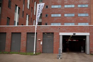 Un bâtiment en briques rouges sur lequel flotte une bannière blanche avec l’inscription « AMSTERDAM HOTEL ». Il y a trois grandes portes de garage et une enseigne au-dessus de l’entrée.