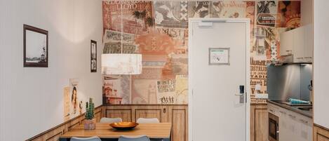 Una cocina compacta con comedor, armarios de madera, un refrigerador de acero inoxidable y una pared decorada con carteles antiguos.