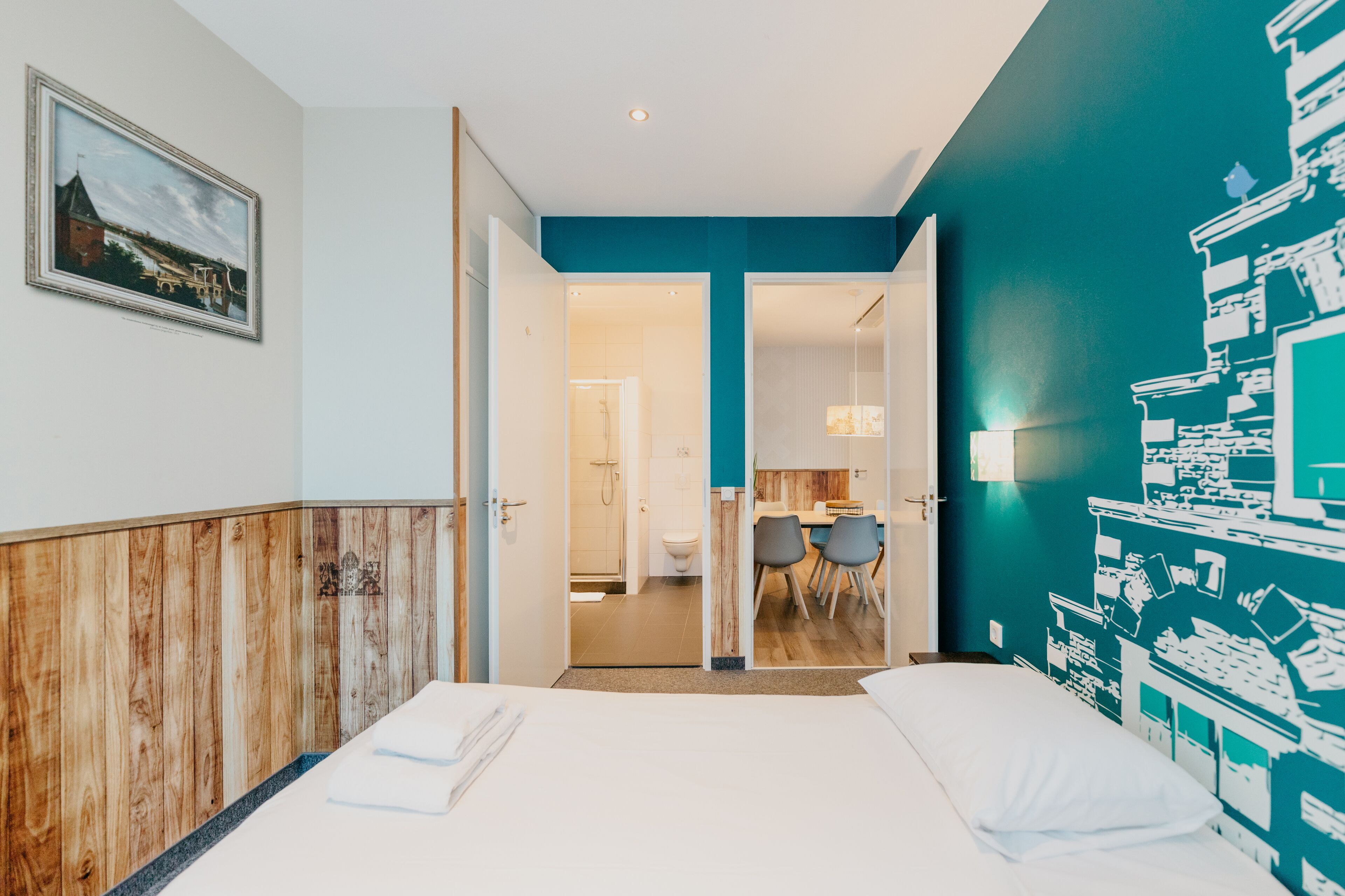 A hotel room with a bed, a wooden panel wall, a teal accent wall, a dining area with chairs, and a painting on the wall.