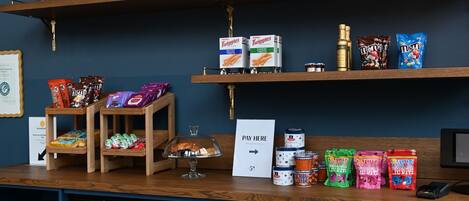 Zona de exhibición de aperitivos y bebidas con diferentes productos envaselados, un letrero que dice 'Pagar Aquí' y un refrigerador lleno de bebidas.