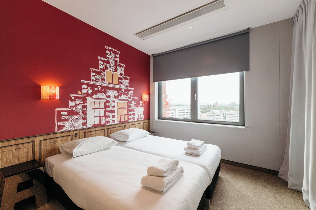 A hotel room with two beds, a red wall with a white abstract design, a large window with a view of buildings, and a ceiling fan.