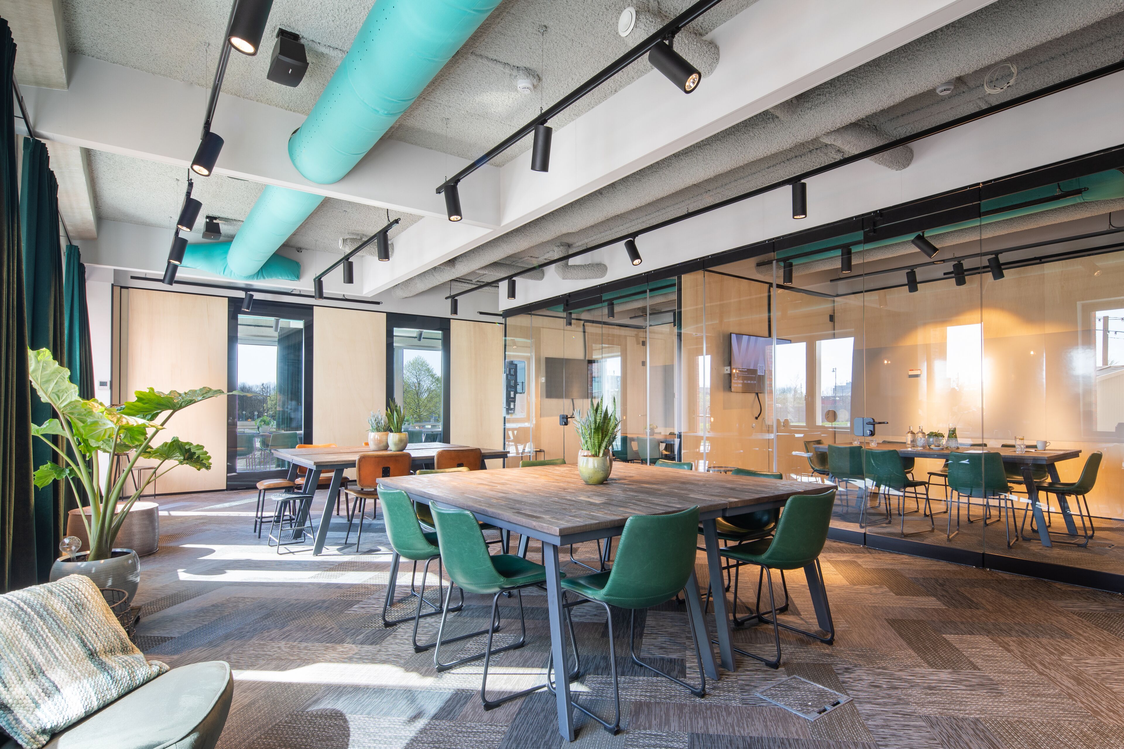 A modern conference room with a large table, green chairs, potted plants, and glass walls.