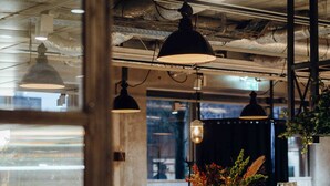 Interior de restaurante con techos altos a la vista, iluminación colgante, una barra de madera y vistas a través de un ventanal.