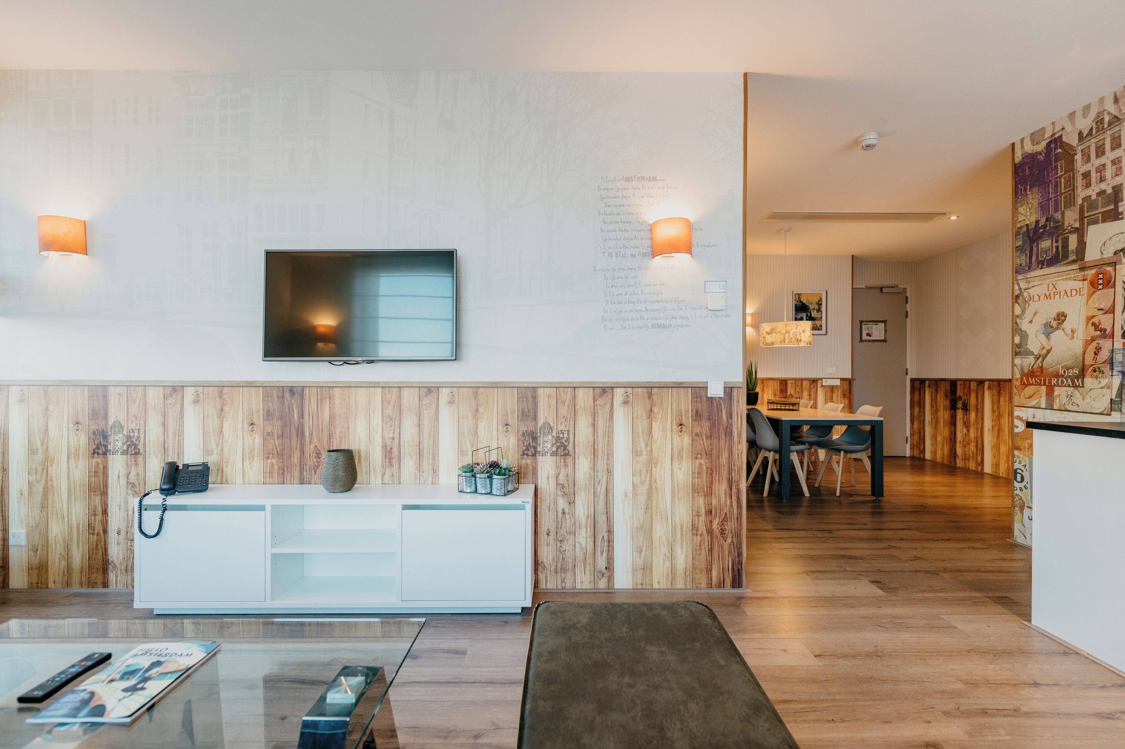 A modern hotel room with a wooden accent wall, a flat-screen TV, a white console, and a dining area with a wooden floor.