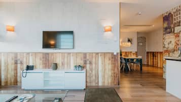 A modern hotel room with a wooden accent wall, a flat-screen TV, a white console, and a dining area with a wooden floor.
