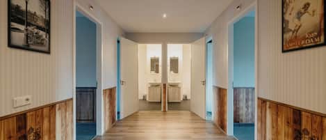 Un couloir d’hôtel avec des lambris en bois, une moquette bleue et une porte miroir donnant sur une salle de bain équipée d’un lavabo et d’un miroir.