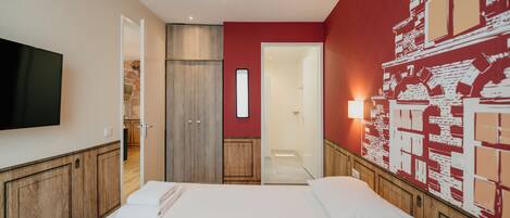 A hotel room with a large bed, a flat-screen TV, wooden cabinets, and a red accent wall with a cityscape mural.