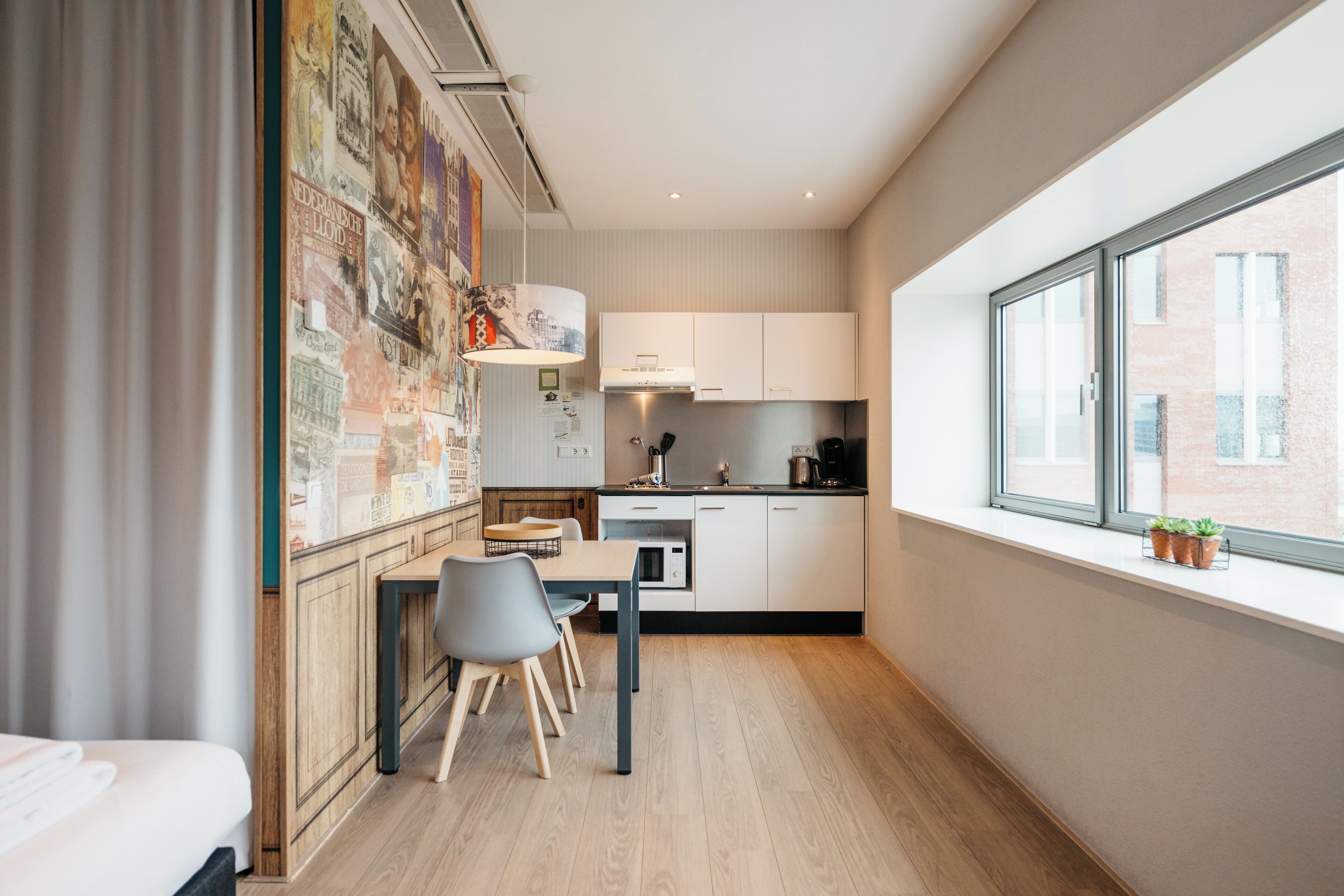 Una habitación de hotel moderna con piso de madera, un área de comedor con mesa y sillas, una kitchenette con armarios blancos y un amplio ventanal con vista a la ciudad.