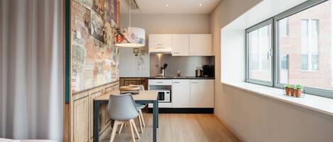 Una habitación de hotel moderna con piso de madera, un área de comedor con mesa y sillas, una kitchenette con armarios blancos y un amplio ventanal con vista a la ciudad.