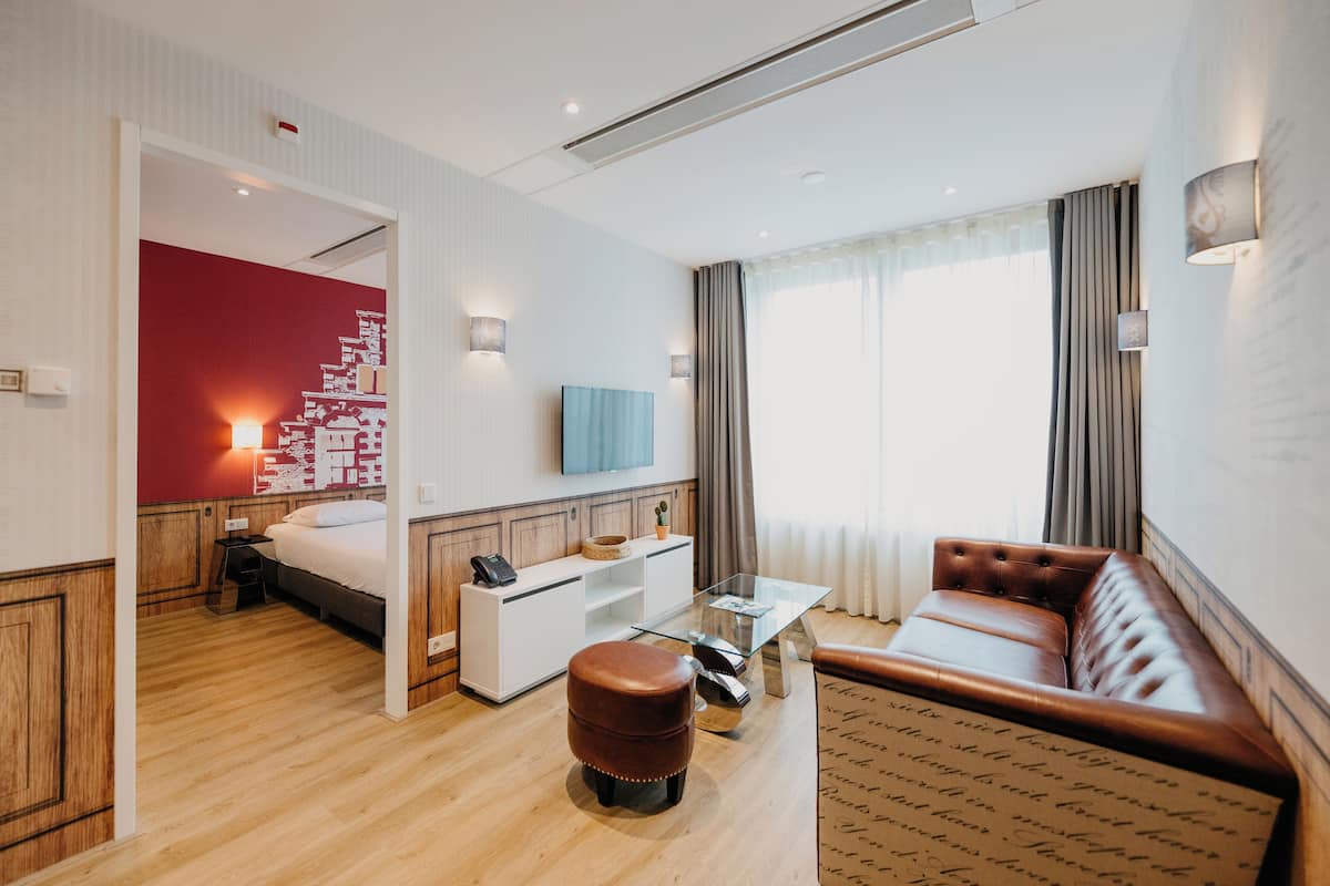 Une chambre d’hôtel avec un plancher en bois, un lit, un canapé, une table basse en verre, un téléviseur et un mur d’accent rouge.