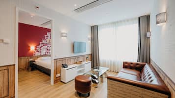 A hotel room with a wooden floor, a bed, a sofa, a glass coffee table, a TV, and a red accent wall.