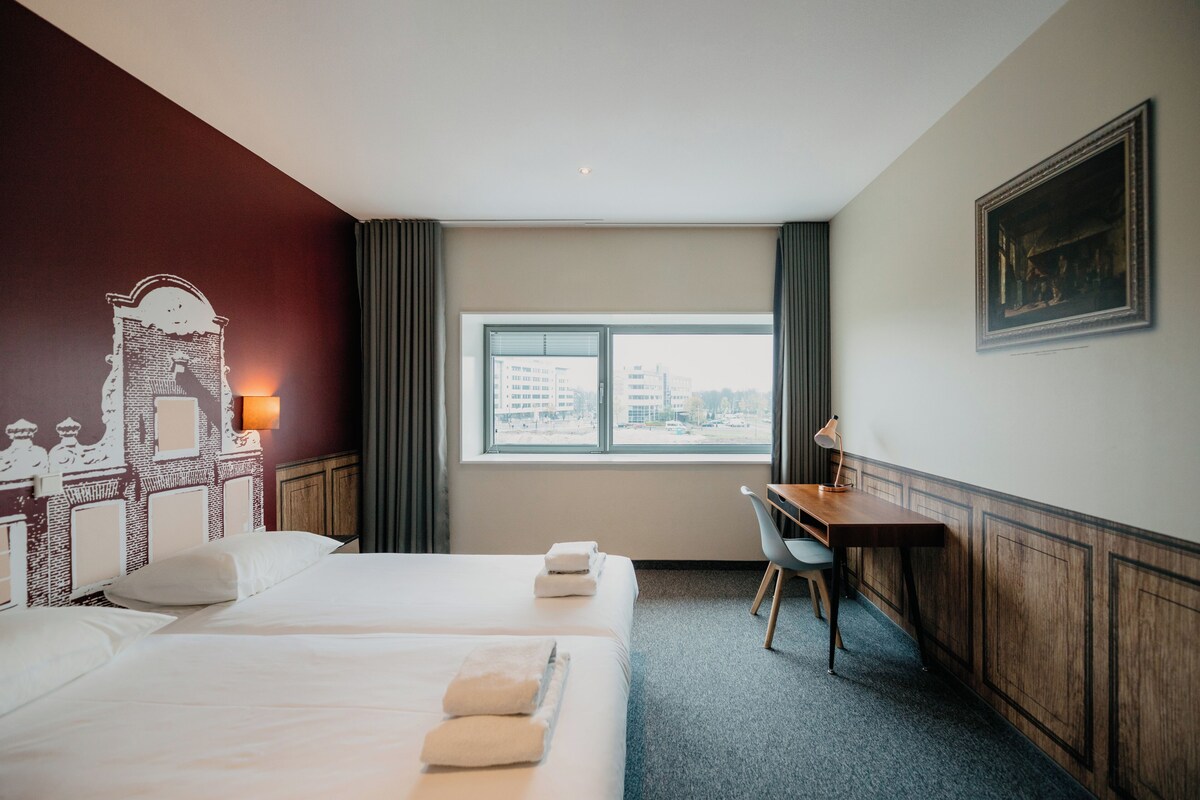 A hotel room with two beds, a desk, a chair, a window with a city view, and a wall-mounted lamp.