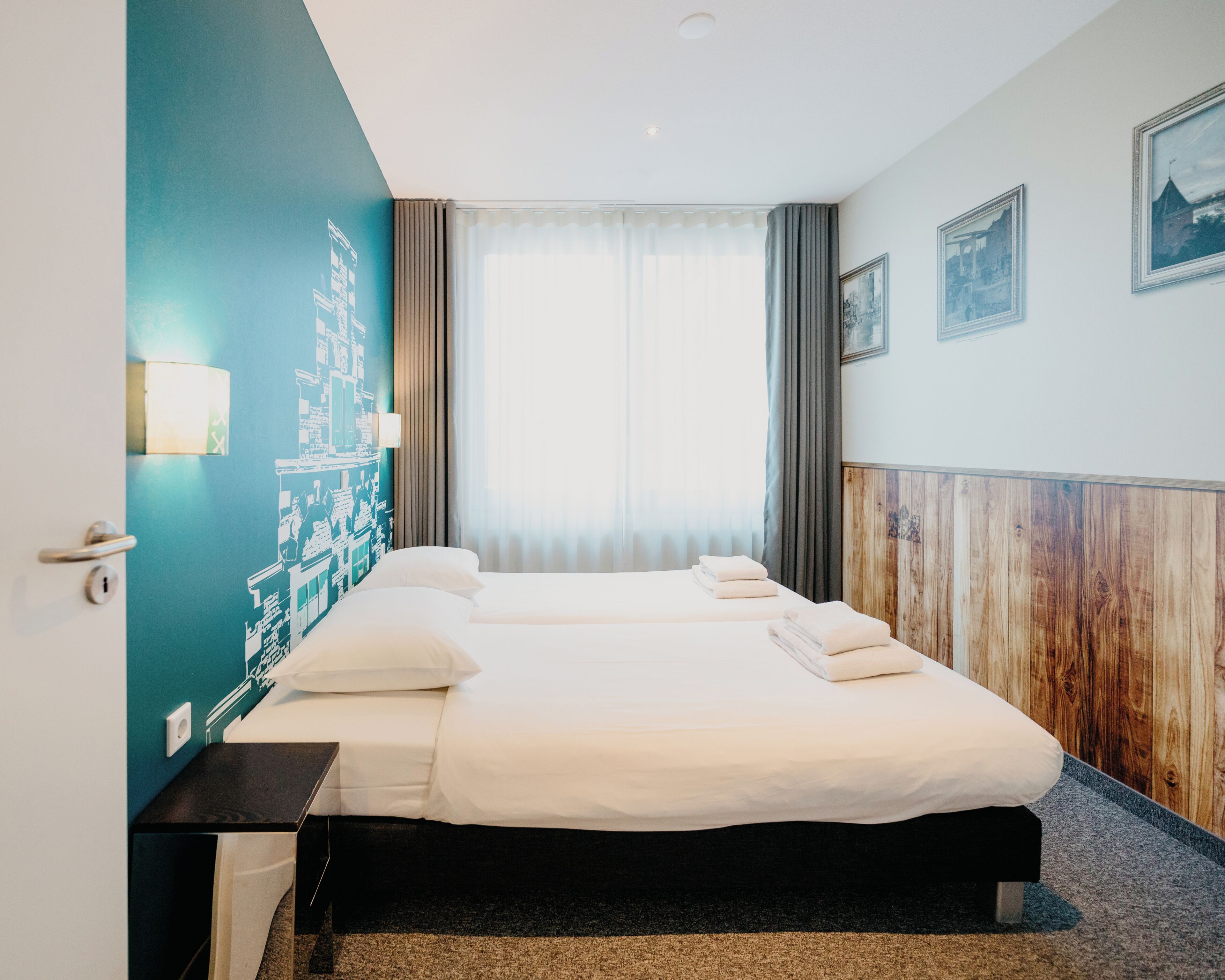 Habitación de hotel con una cama grande, una mesita de noche, una lámpara de pared y un ventanal con cortinas.