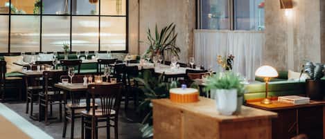 A restaurant with multiple dining tables and chairs, potted plants, and a green sofa in the seating area.