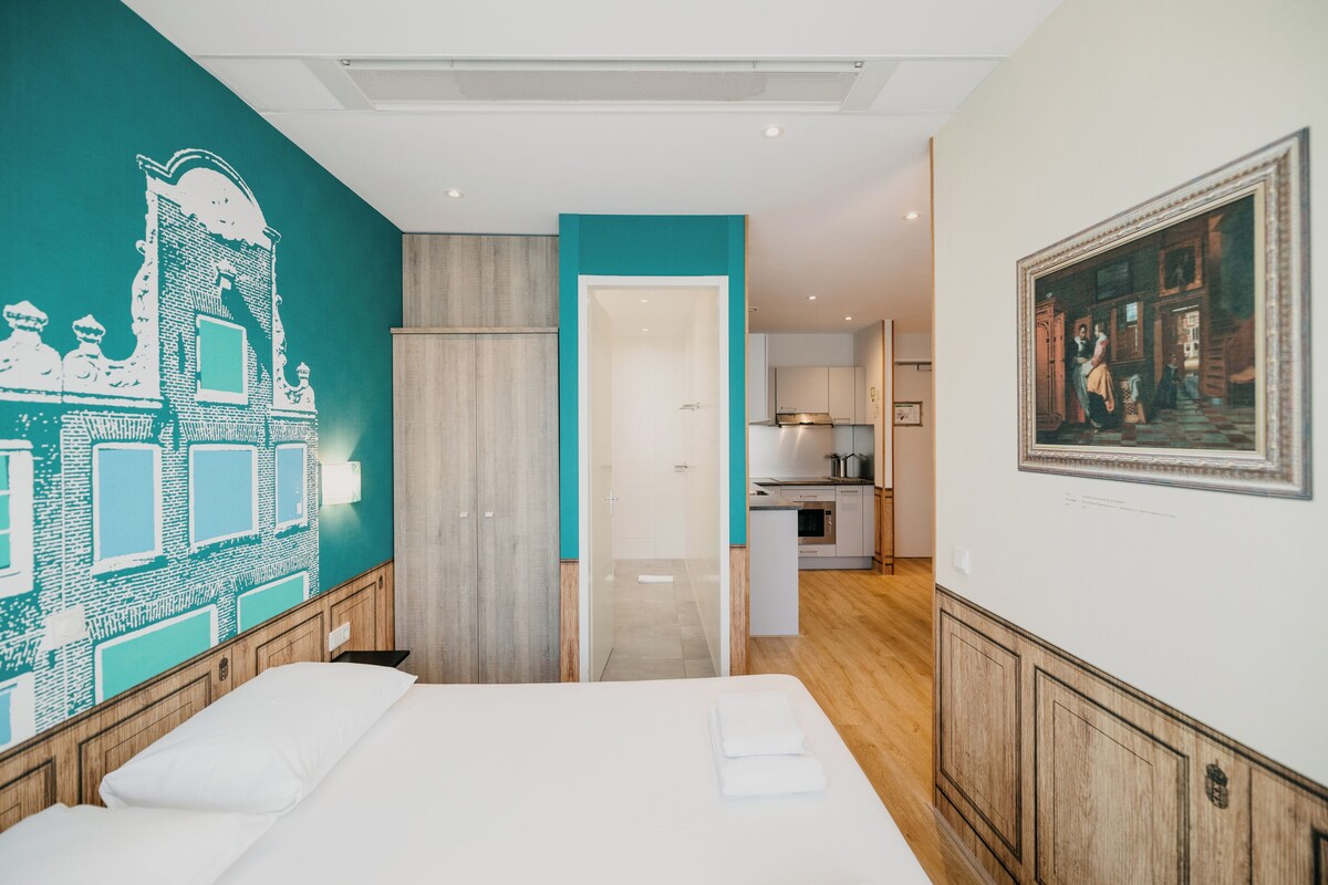 A hotel room with a bed, a teal accent wall, a wooden door, a kitchen area, and a framed painting on the wall.
