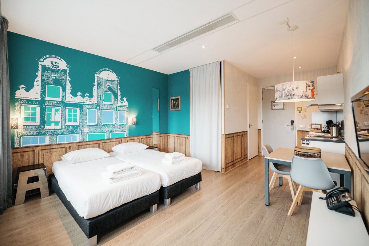 A hotel room with two beds, a desk, and a kitchenette.