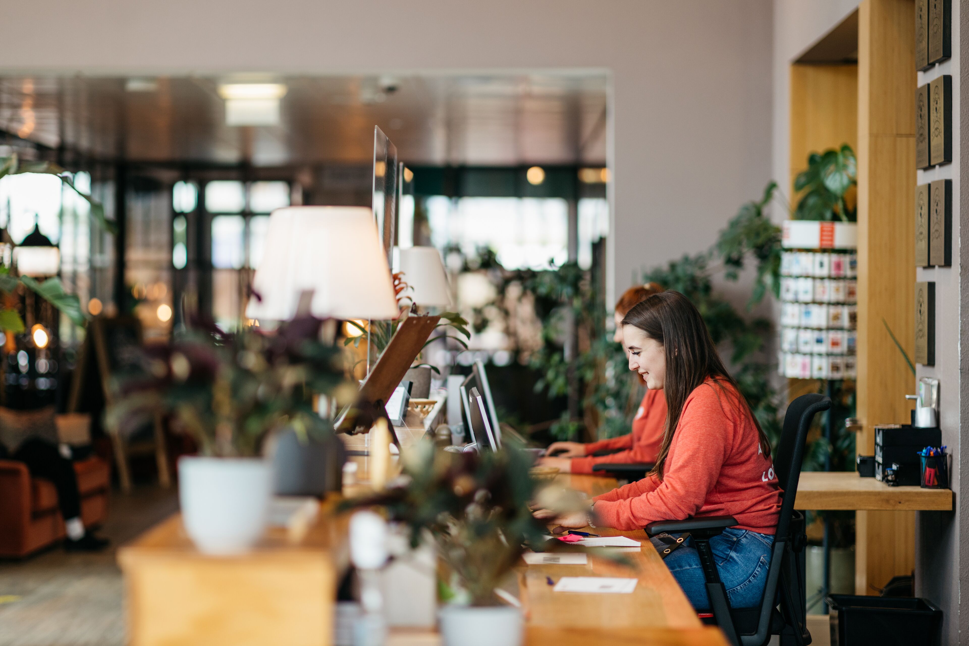 A person working at a desk in a well-lit indoor space with plants and shelves in the background.