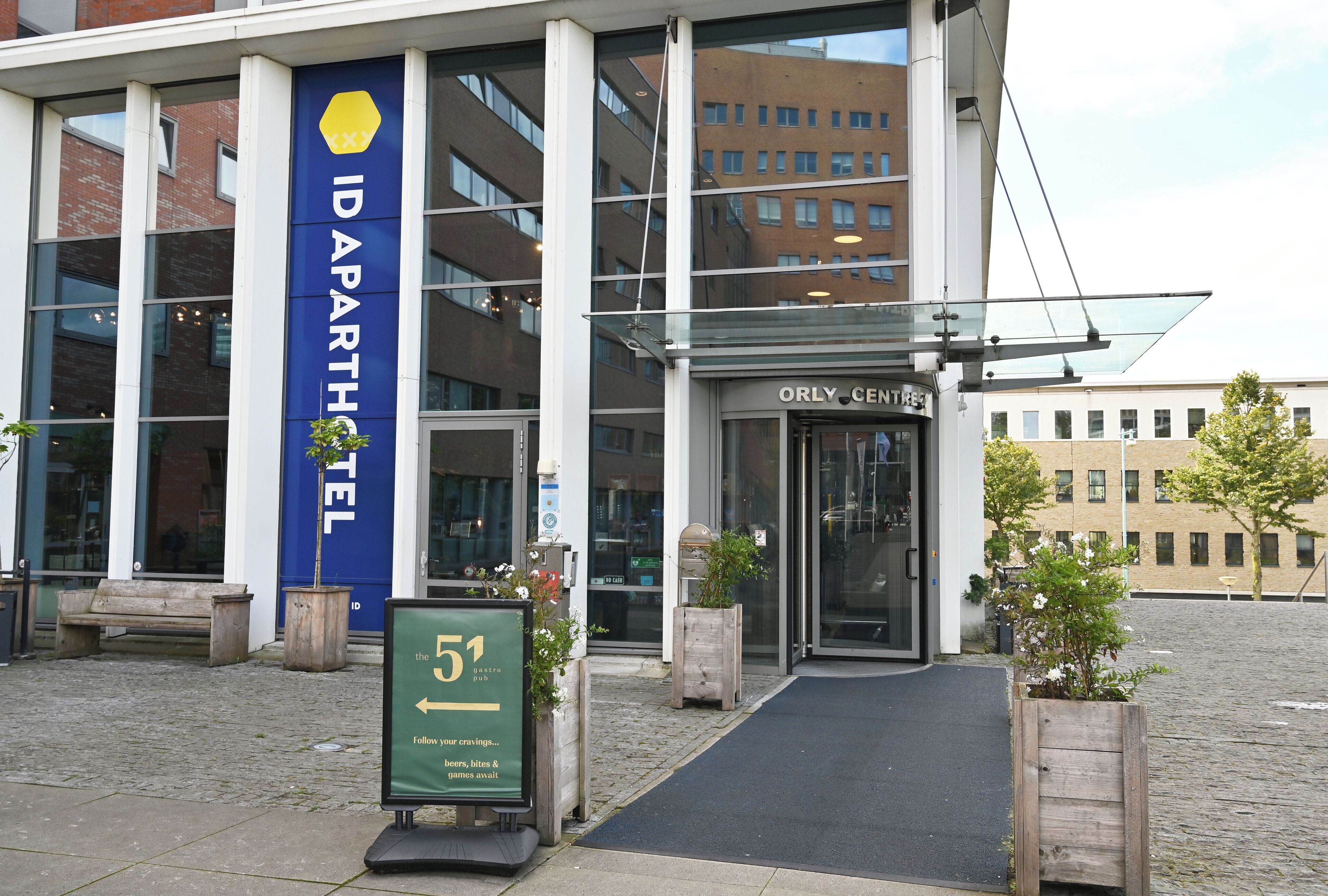 The IDAPARTHETEL hotel entrance with a clear glass facade, a signboard, and a wooden bench outside.