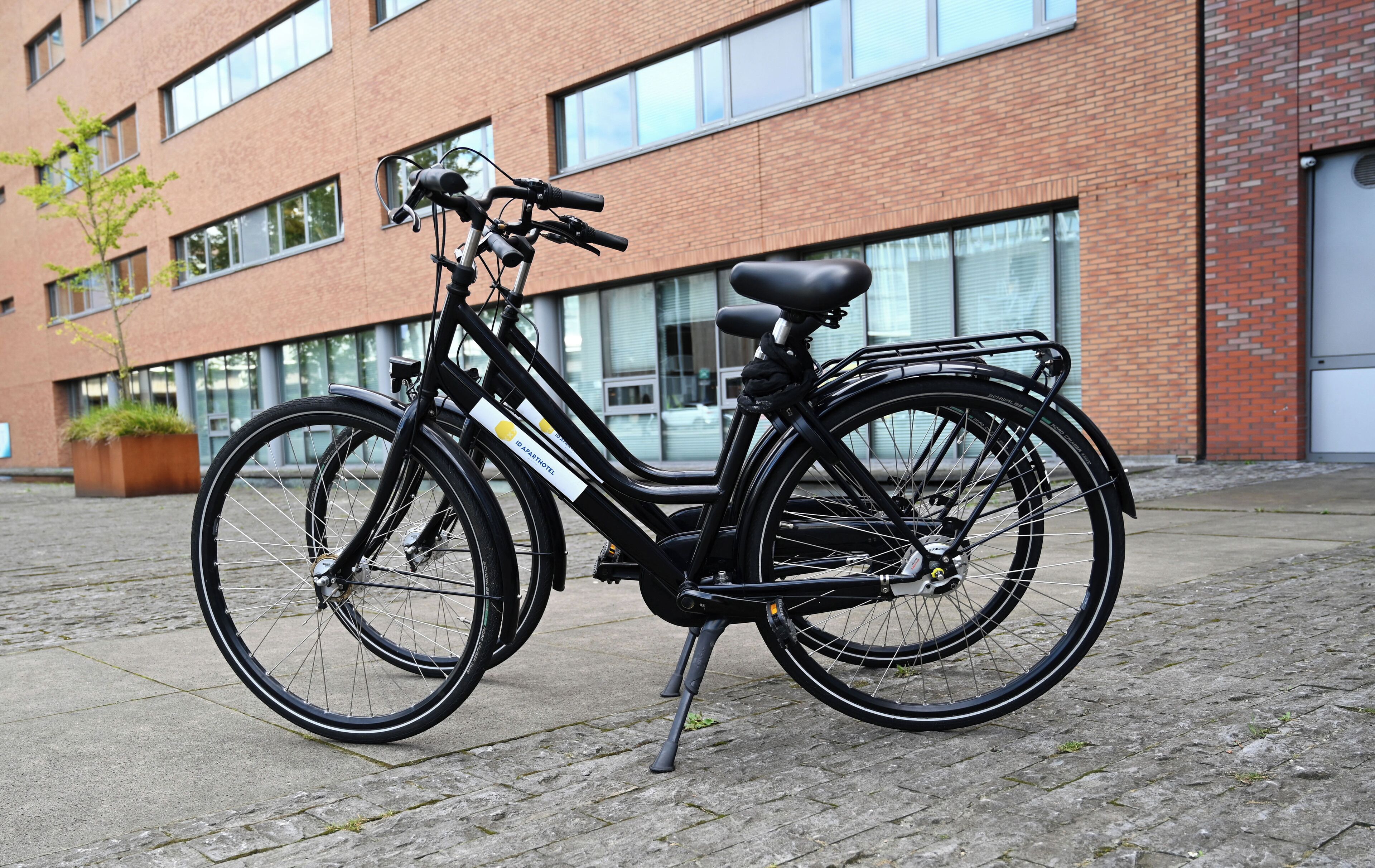A black bicycle with a rear rack parked on a cobblestone path in front of a brick building with large windows.
