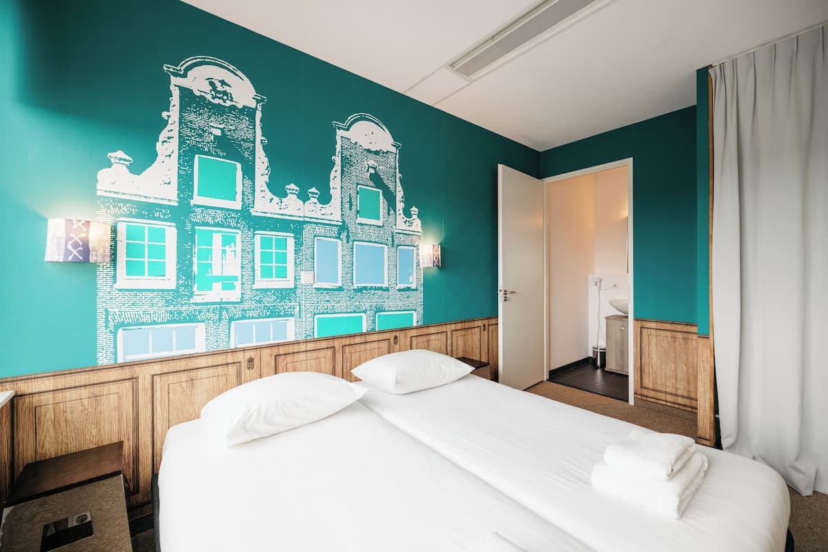 Une chambre d’hôtel avec un grand lit, une tête de lit en bois et un mur de couleur turquoise orné d’une fresque architecturale blanche.
