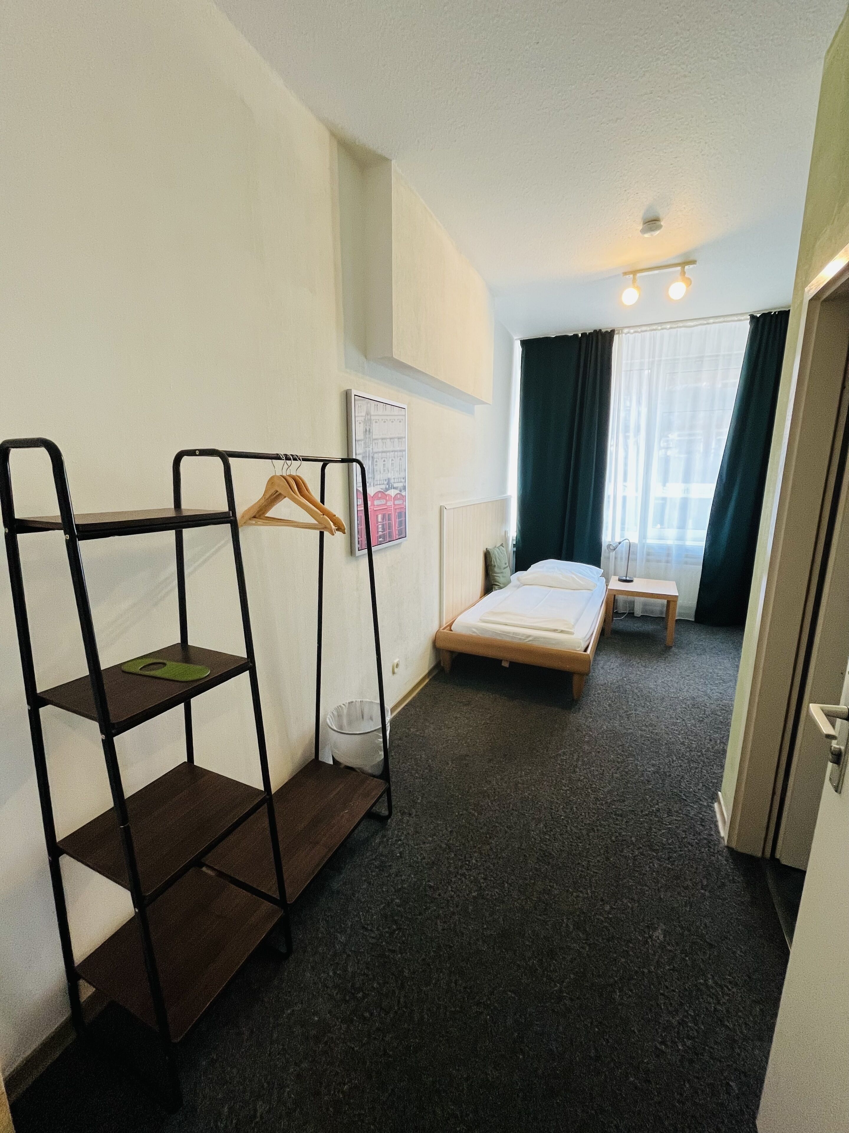 A small hotel room with a bunk bed, a single bed, a wooden bench, a coat rack, a small table, and a wall-mounted picture.