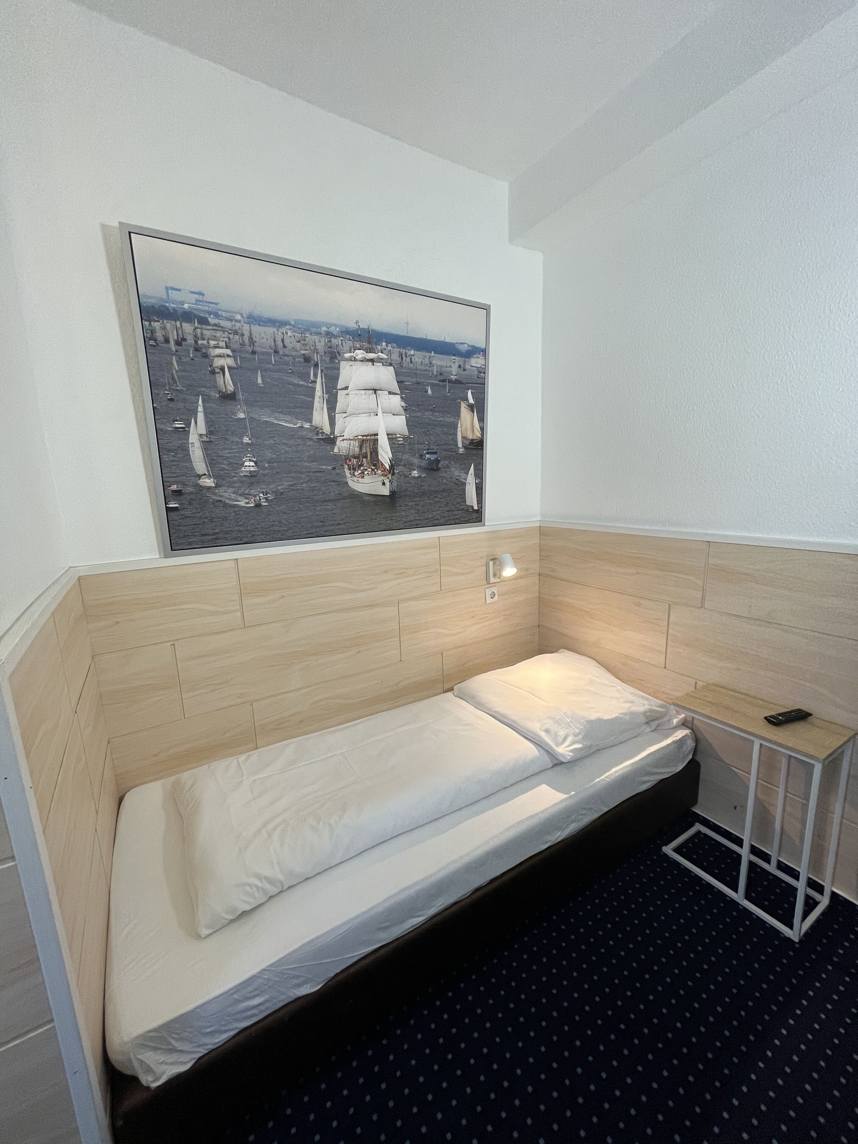 A small, single-bed room with a wooden headboard, a bedside table with a lamp, and a large framed picture of a sailing scene on the wall.