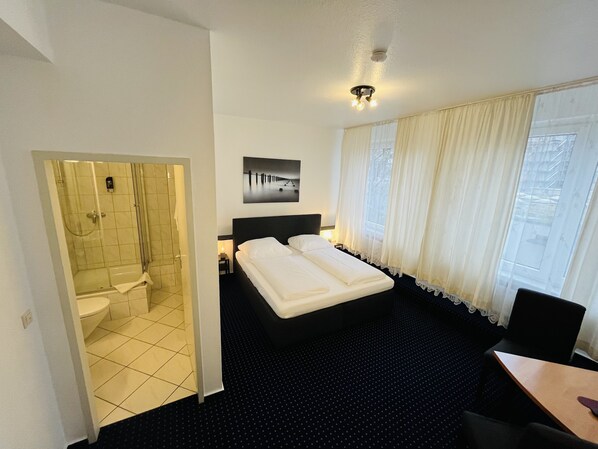 A hotel room with a bed, a desk, a chair, a bathroom with a shower and toilet, and a window with curtains.