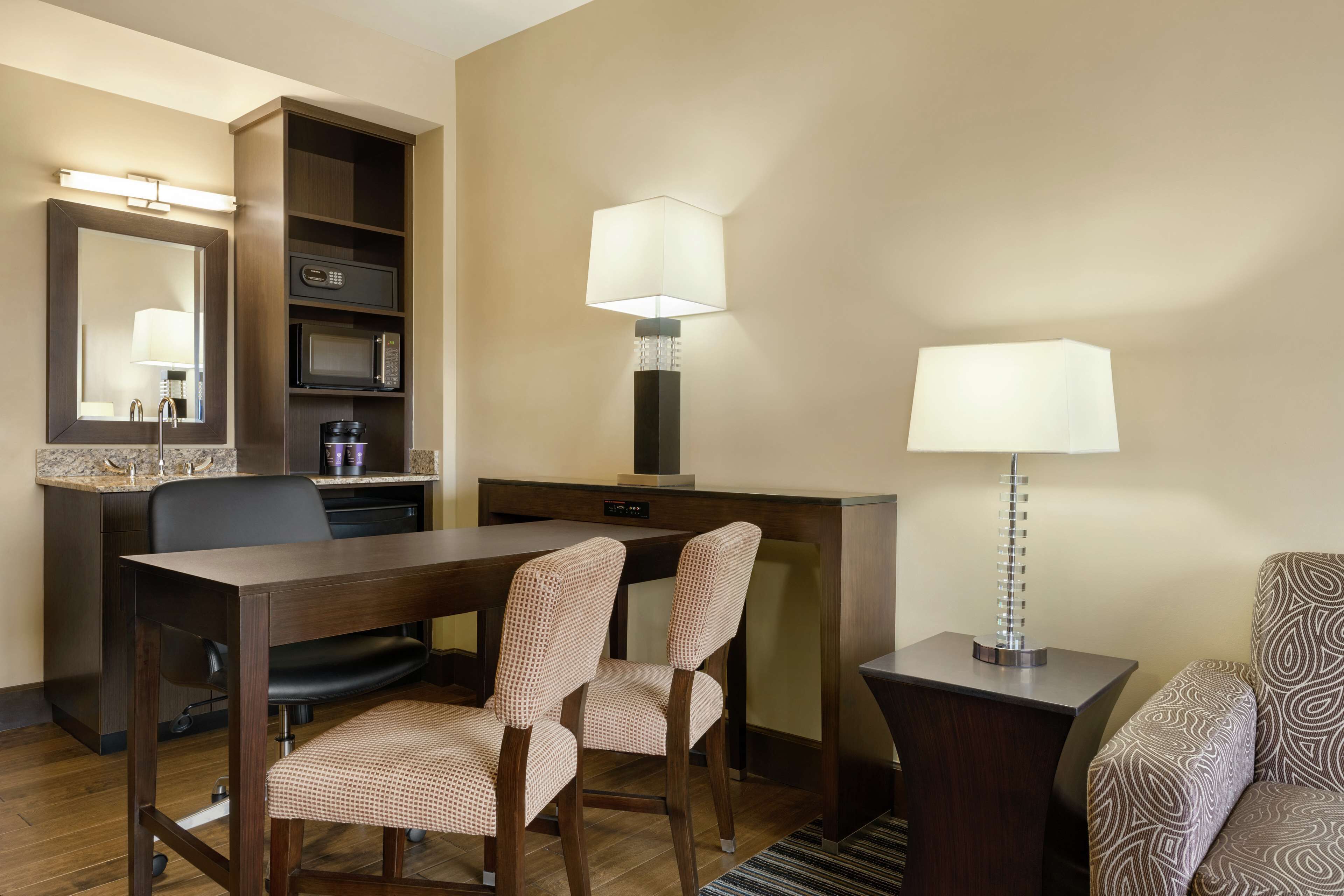 A hotel room with a desk, two chairs, a microwave, a coffee maker, a lamp, and a sofa.