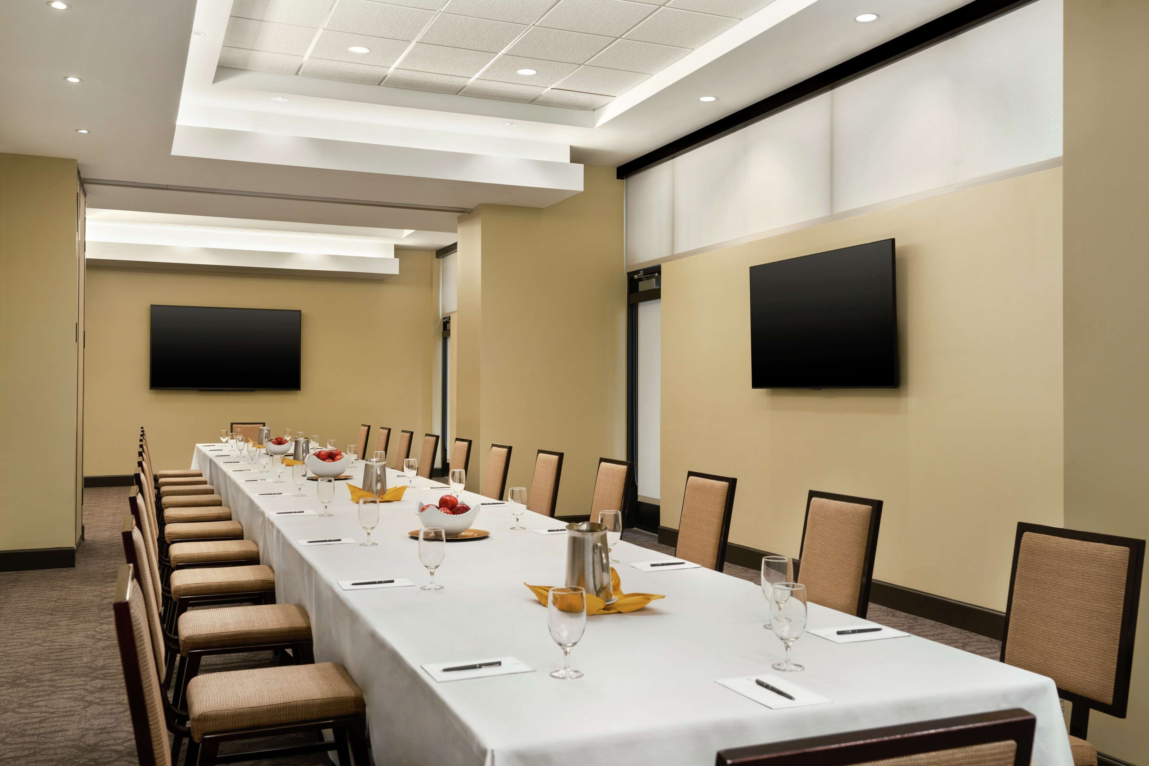 A conference room with a long table set for a meeting, equipped with chairs, a projector screen, and a flat-screen TV.